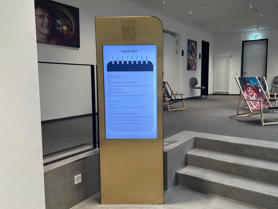 Foto eines digitalen Infoscreens in einem modernen Innenraum mit Veranstaltungskalender, umgeben von Stühlen, Bildern an den Wänden und Liegestühlen mit bunten Motiven.